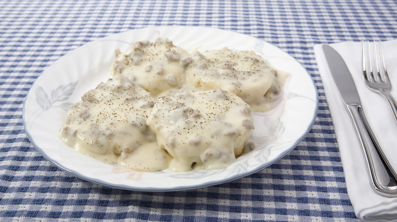 biscuits drowning in white gravy on a white plate sitting on a table with a blue and white gingham tablecloth next to a napkin, butter knife, and fork