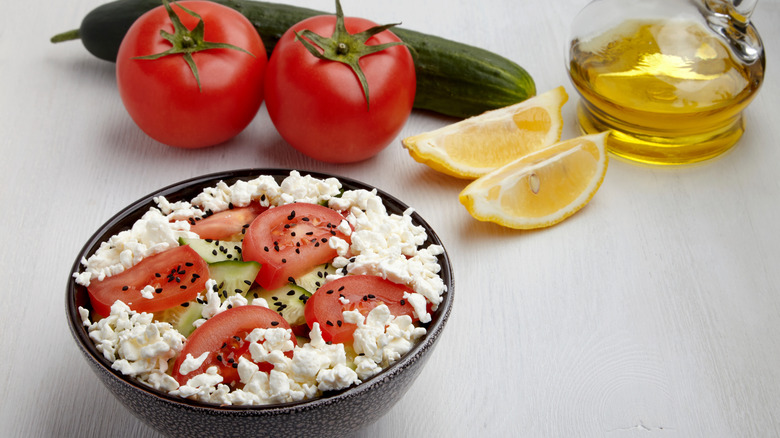 Bowl of cottage cheese with tomatoes and cucumbers with seasoning and veggies in background with lemon slices next to olive oil