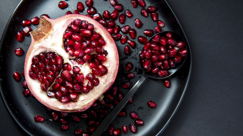 Split pomegranate on black plate next to spoon holding arils