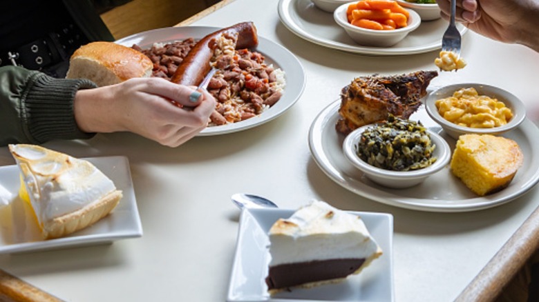 Two people eating a Southern food meal with plates of gumbo, chicken with collards and mac and cheese, cornbread, lemon meringue pie, and chocolate meringue pie