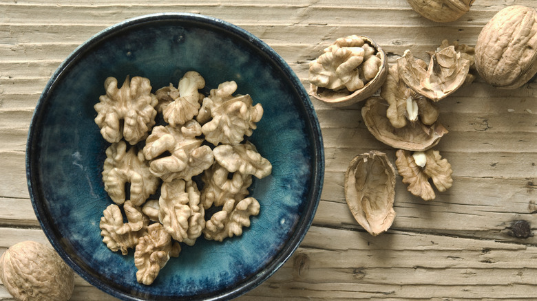 A bowl of shelled walnuts, surrounded by whole walnuts on a rustic wooden table