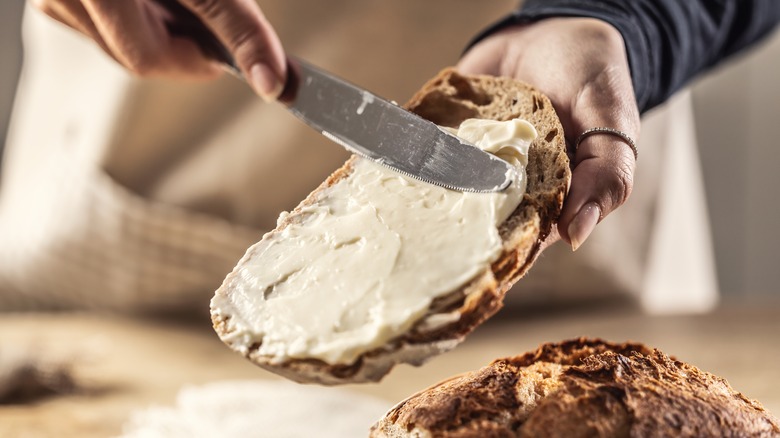 A person spreads cream cheese on a slice of toast, on a rustic wooden bread paddle