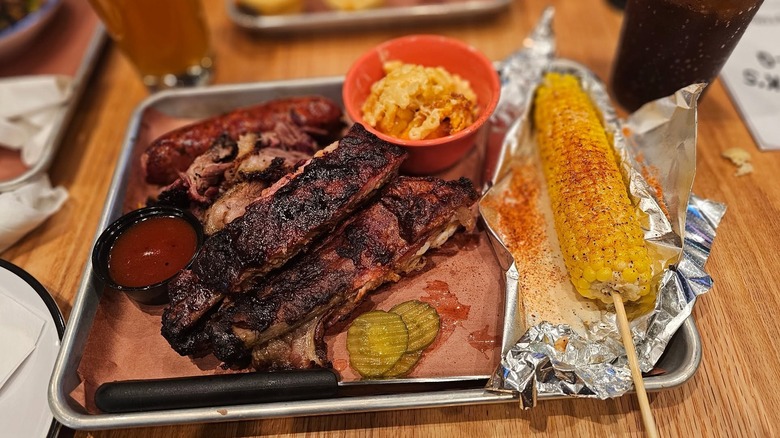 A BBQ rib platter with sausage, corn, and macaroni and cheese on it with pickles and sauce