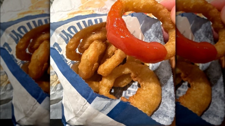 Hand holding Culver's onion ring dipped in ketchup over bag of onion rings