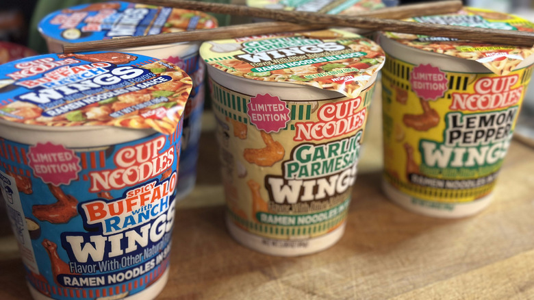 Three new Cup Noodles flavors lined up on table with chopsticks, including spicy buffalo with ranch wings, garlic parmesan wings, and lemon pepper wings