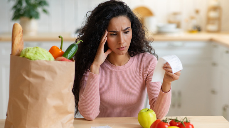 Woman reading receipt looking confused near bag of groceries in kitchen