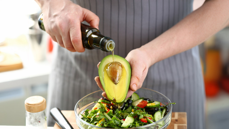 A person pouring oil over half an avocado above a bowl of salad.