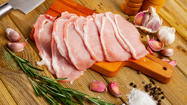 Raw thin pork chops on wooden surface with fresh rosemary and garlic cloves