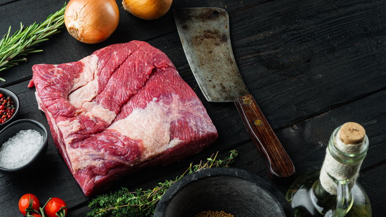 A whole piece of raw Beef Brisket with a cleaver, onions, fresh herbs, salt, and peppercorns