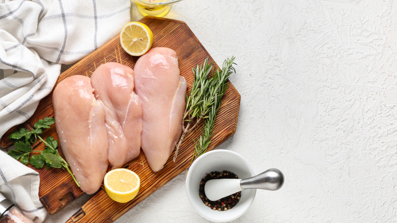 Wooden board with raw chicken breast, fresh herbs, and lemon halves
