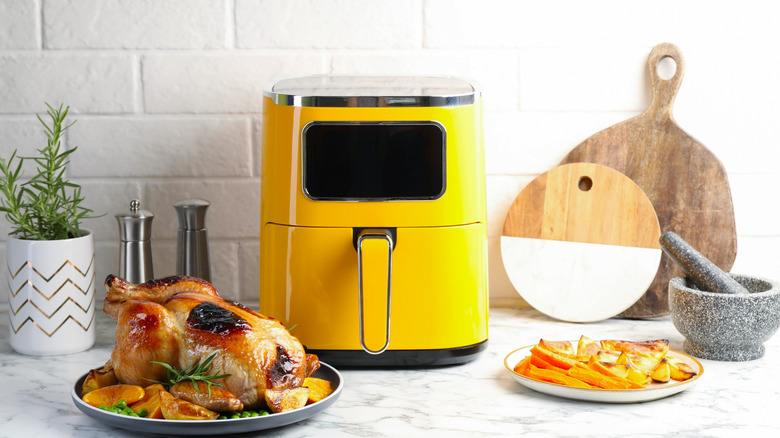 Roasted chicken on a plate, sweet potato fries on a plate, yellow air fryer, mortar and pestle, salt and pepper grinders, rosemary plant in a white vase, and two wooden paddles in the background
