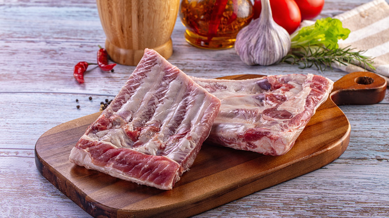 Raw pork ribs on a wooden cutting board with fresh vegetables, a head of garlic, and herbs in the background