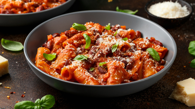 A grey bowl of pasta e fagioli garnished with basil leaves and grated parmesan cheese