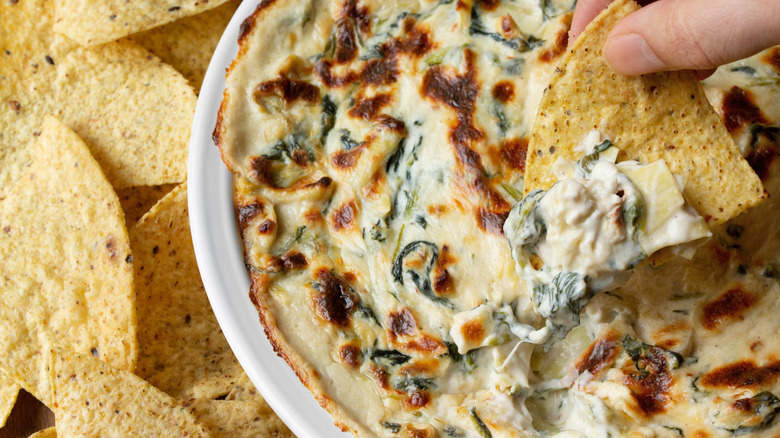 White bowl of cheesy spinach and artichoke dip surrounded by tortilla chips