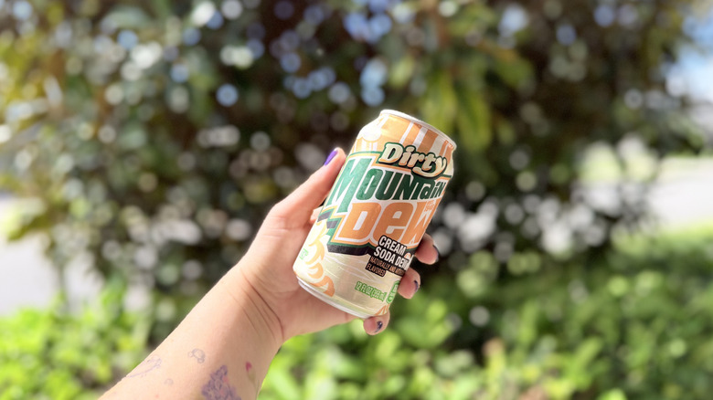 Holding can of mountain dew dirty cream soda on blurred background of greenery