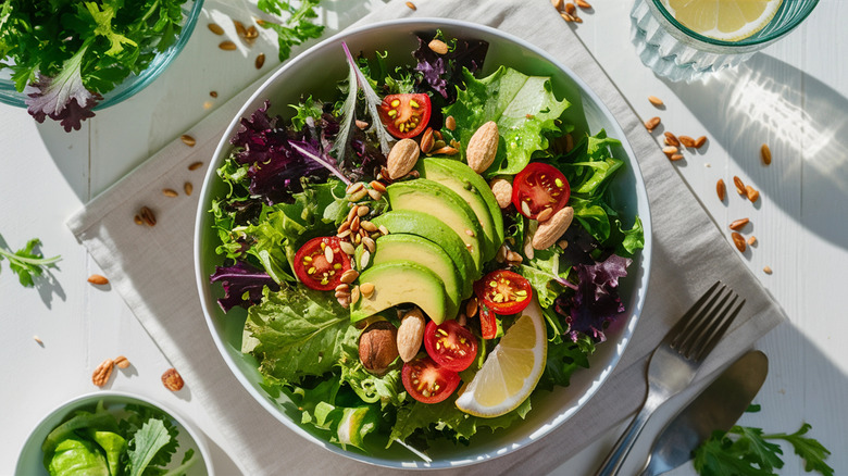 White bowl with green salad with avocado, tomato, nuts, and lemon wedge