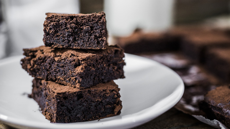 A close up of three brownie squares stacked on a white plate, with a jar of milk out of focus in the background