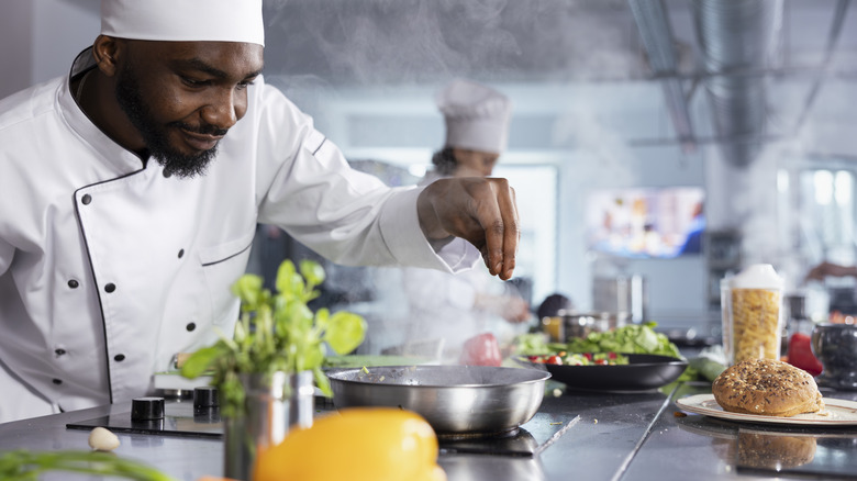 A chef seasons a dish over a fry pan in a kitchen