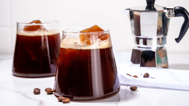 Two glass cups of coffee with espresso ice cubes in them and a steel coffee pot behind them
