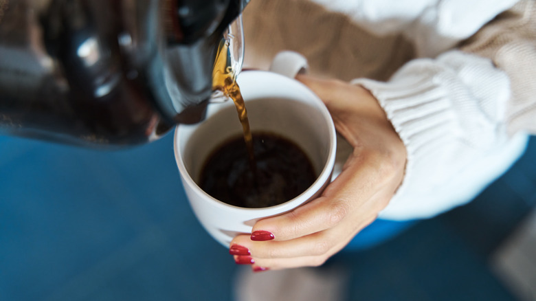A woman's hand holds a coffee cup into which coffee is poured from a carafe