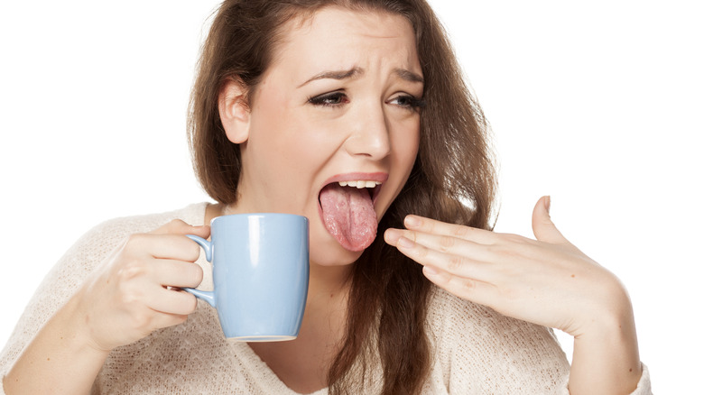 A woman, wearing a pained expression, sticks out her tongue while holding a coffee mug