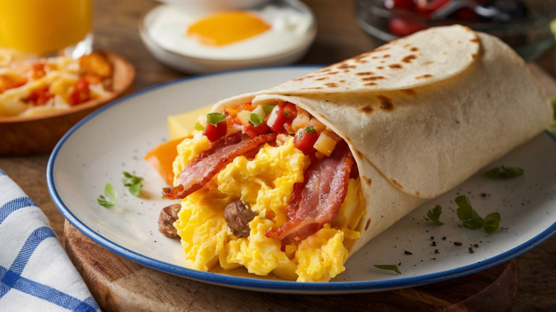 Breakfast burrito with eggs, bacon and veggies on round white plate with a blue border on a round wood board