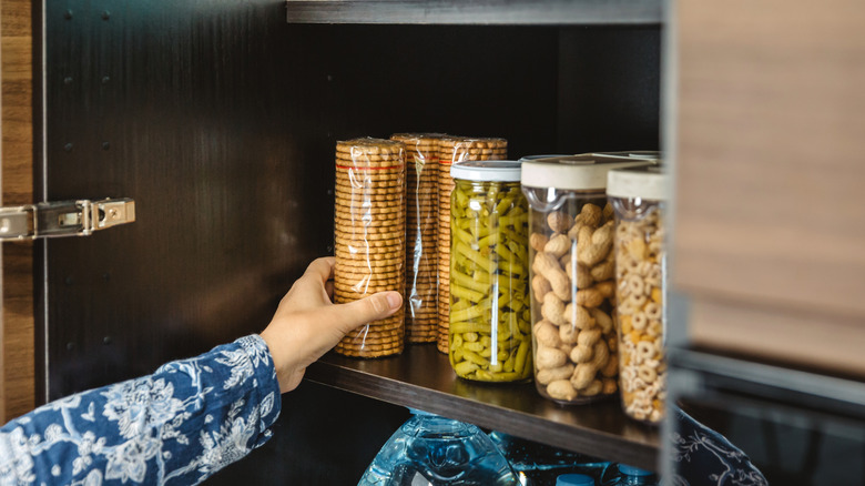 A hand reaching into a cupboard where food is stored in glass jars and crackers are in plastic sleeves