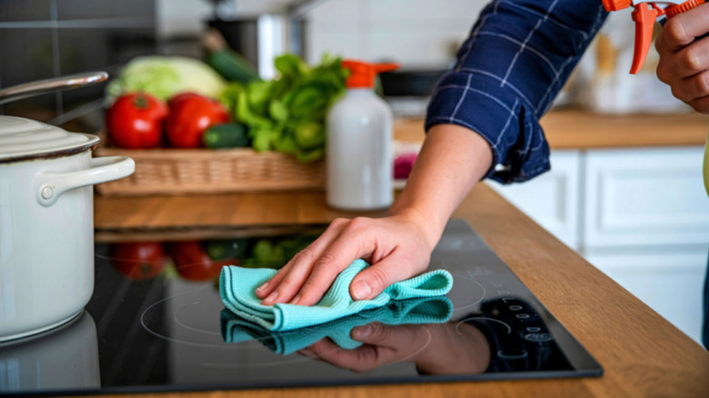 A hand wipes a clean glass stovetop with a blue cloth and spray cleaner.
