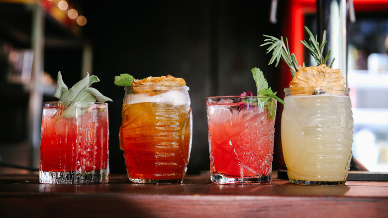 A line of cocktails with fruit and herb garnishes sitting on a bar counter.