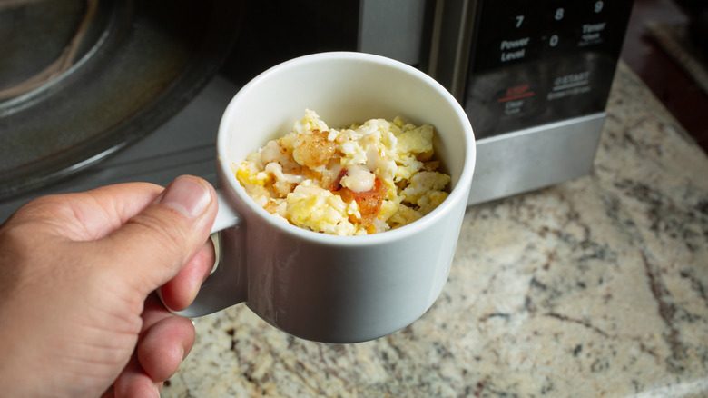 A hand holding a mug of scrambled eggs in front of a microwave