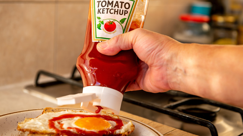 A hand squeezing a bottle of Heinz tomato ketchup on to fried eggs on toast.