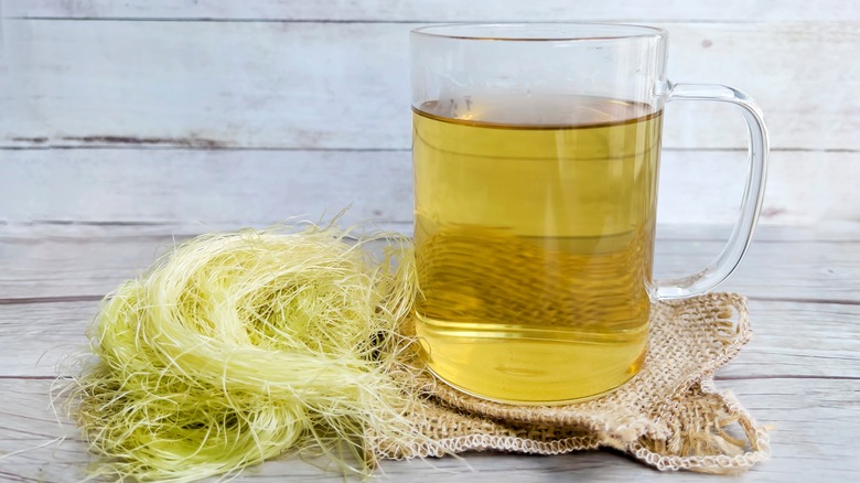 A glass mug of corn silk tea with raw corn silk next to it