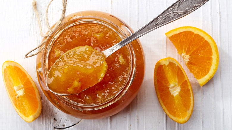 Looking down into a jar of orange marmalade with a full spoon held over it and three orange slices around it on a white surface