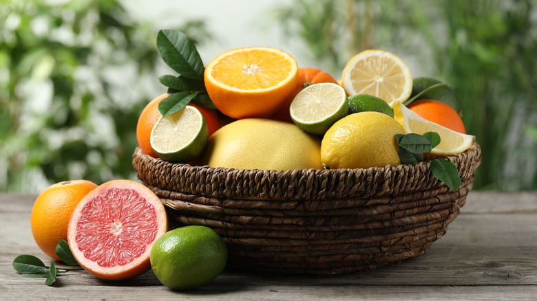 Orange, yellow, and green citrus fruit, whole and cut in half, in and on the side of a brown basket on a wood surface