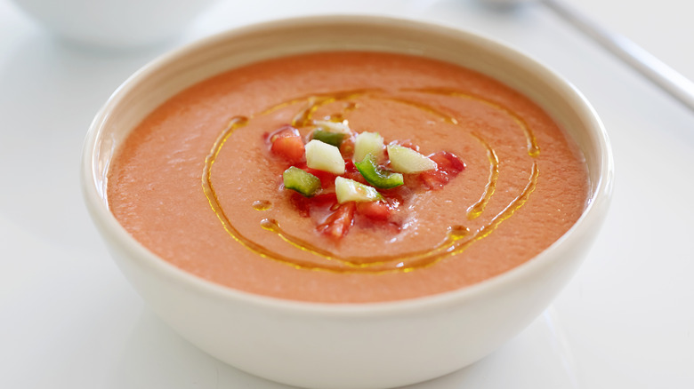 A white bowl of gazpacho with fresh vegetables and a drizzle of oil
