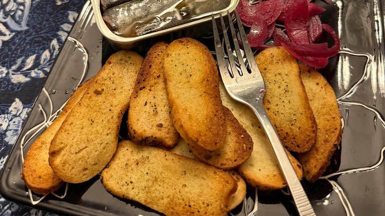 A stack of bagel chips on a plate with a fork on top alongside pickled onion and canned sardines