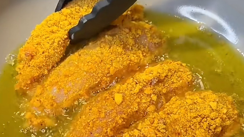 Four chicken tenders coated in everything bagel breadcrumbs are frying in a pan