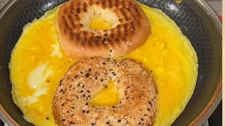 Two halves of a bagel are being cooked in a pan facedown, on top of a layer of scrambled eggs