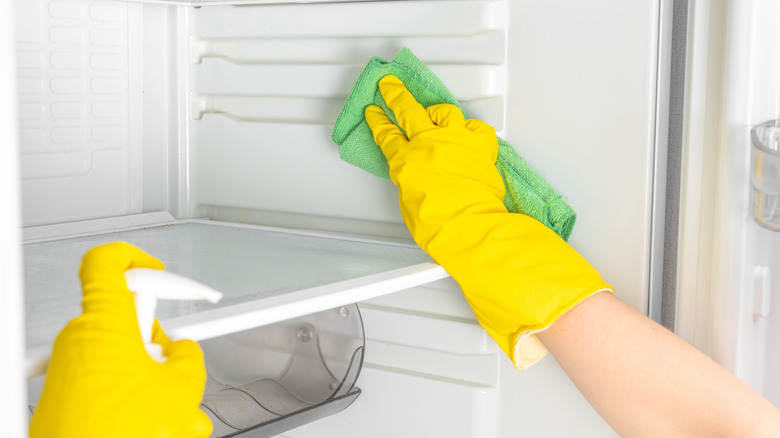 A person wearing rubber gloves with cleaner and a towel wiping the inside of a fridge