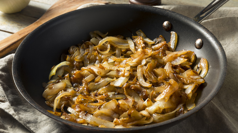 Caramelized onions sitting in a frying pan on a table with a cloth and wooden spoon nearby.