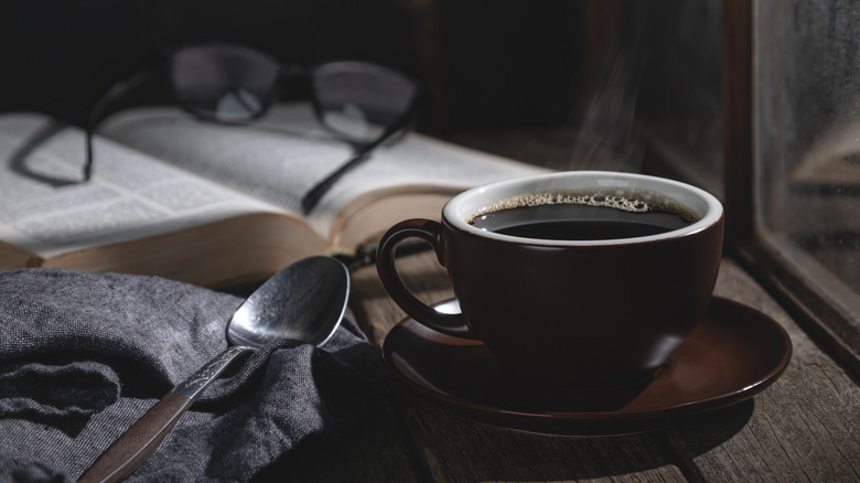 Cup of coffee on a wooden table in dim lighting with book and glasses in the background