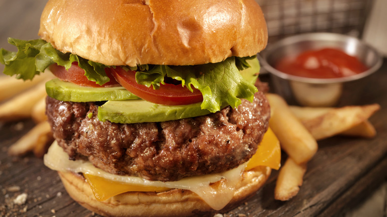 Closeup of a cheeseburger with lettuce, tomato, avocado, and a side of fries.