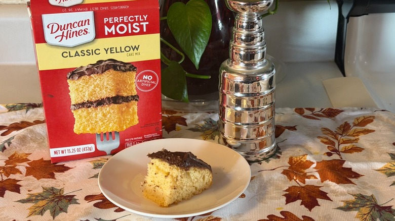 A piece of Duncan Hines yellow cake with chocolate frosting on a plate in front of the box and a small replica of the Stanley Cup.