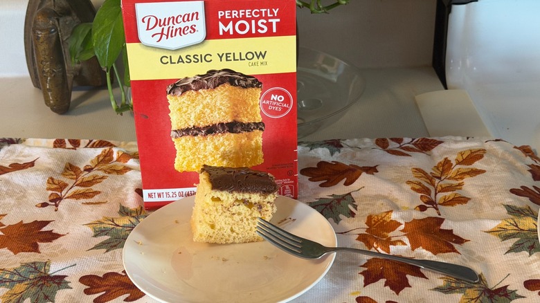 A piece of Duncan Hines yellow cake with chocolate frosting on a plate with a fork in front of the box.