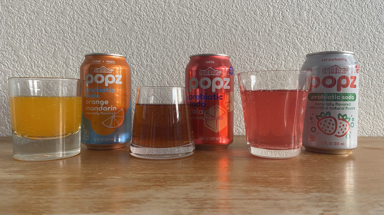 Popz cans arranged horizontally beside glasses full of each soda