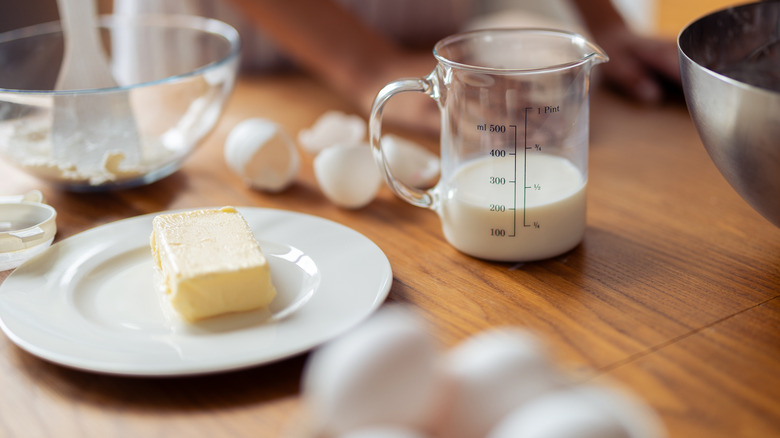Kitchen scene with milk, butter, eggs, and mixing bowls on a wooden table, with people blurred in the background.