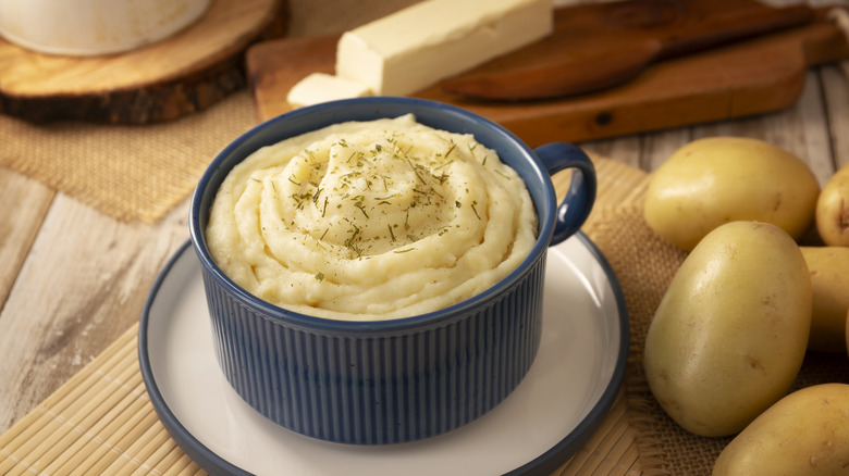 A blue bowl of smooth mashed potatoes with butter and potatoes nearby.