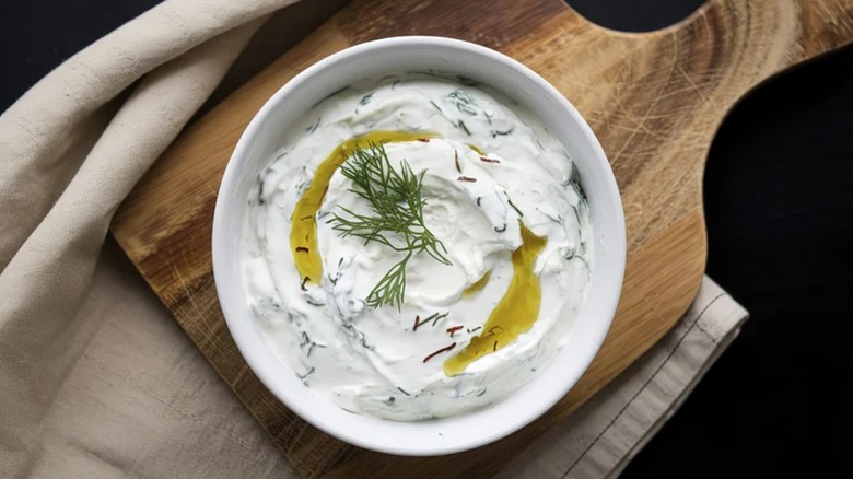 A white bowl of Greek yogurt dip topped with fresh dill sitting on a wooden cutting board
