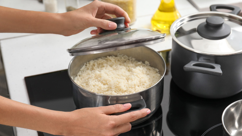 Woman holds pot handle and removes lid from saucepan of cooked white rice on a stove