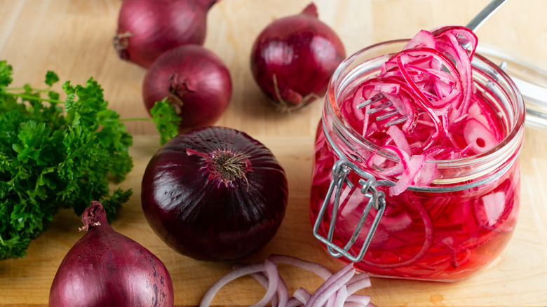 Jar of pickled red onions on a wooden surface surrounded by onions.